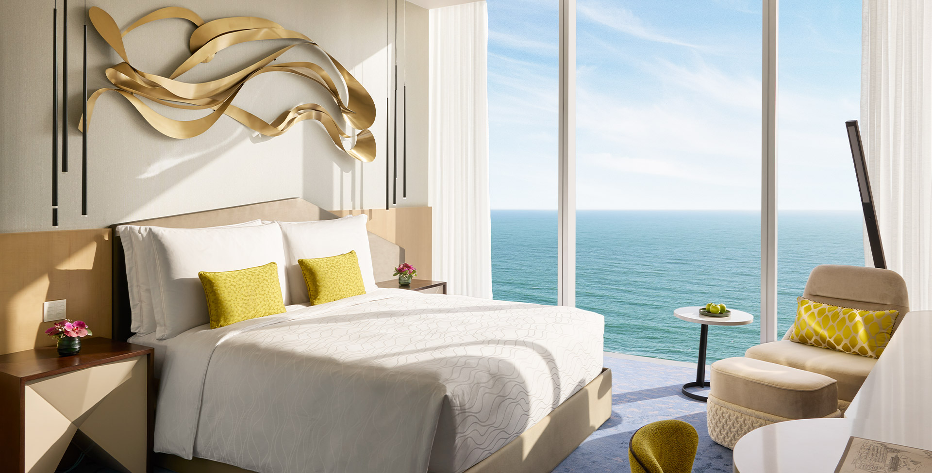 A Deluxe King Oceanfront room, a king bed under a gold wall sculpture and a wall of glass over the sea